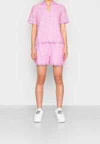 Top in pizzo rosa con maniche corte, orlo scallop e pantaloni corti abbinati. Indossato con sneaker bianche con accenti colorati.