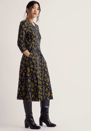 Woman standing wearing a navy blue dress with yellow floral print and black knee-high heeled boots, hands in pockets, looking forward.