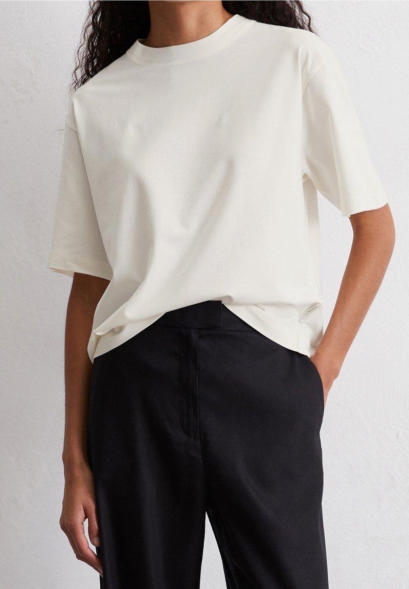 Femme portant un t-shirt blanc uni à manches courtes, légèrement rentré dans un pantalon noir taille haute, une main dans la poche.