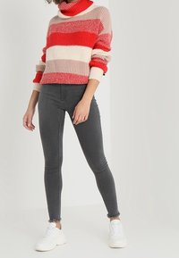 Pull épais à rayures rouges, roses et crème avec un col montant, associé à un jean skinny gris foncé et des baskets blanches.