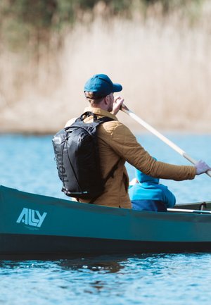 Zwarte rugzak, gewatteerde tan jas, houten peddel, blauwe pet, persoon die op het water kanoet, met een andere persoon in een blauwe hoodie. "ALLY" logo zichtbaar.