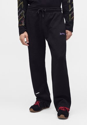 Personne portant un pantalon de jogging noir avec taille à cordon et petits logos brodés, associé à des baskets noires avec des lacets rouges.