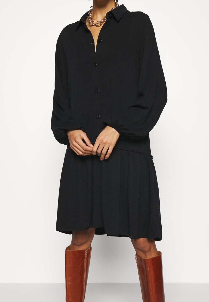 Robe noire boutonnée avec col, manches ballon et jupe plissée. Accompagnée de longues bottes en cuir marron et d'un collier chaîne argenté.