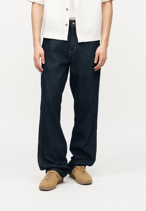 Oseba, oblečena v široke temno modre jeans hlače, rjave semiš superge in belo srajco s kratkimi rokavi in gumbi, stoji na belem ozadju.