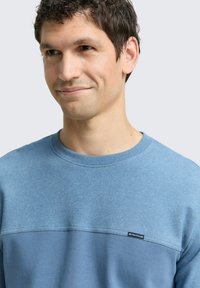Niebieska bluza wykonana z miękkiego materiału, cechująca się dwukolorowym wzornictwem, okrągłym dekoltem oraz subtelną naszywką z logo z przodu. Gładka faktura.