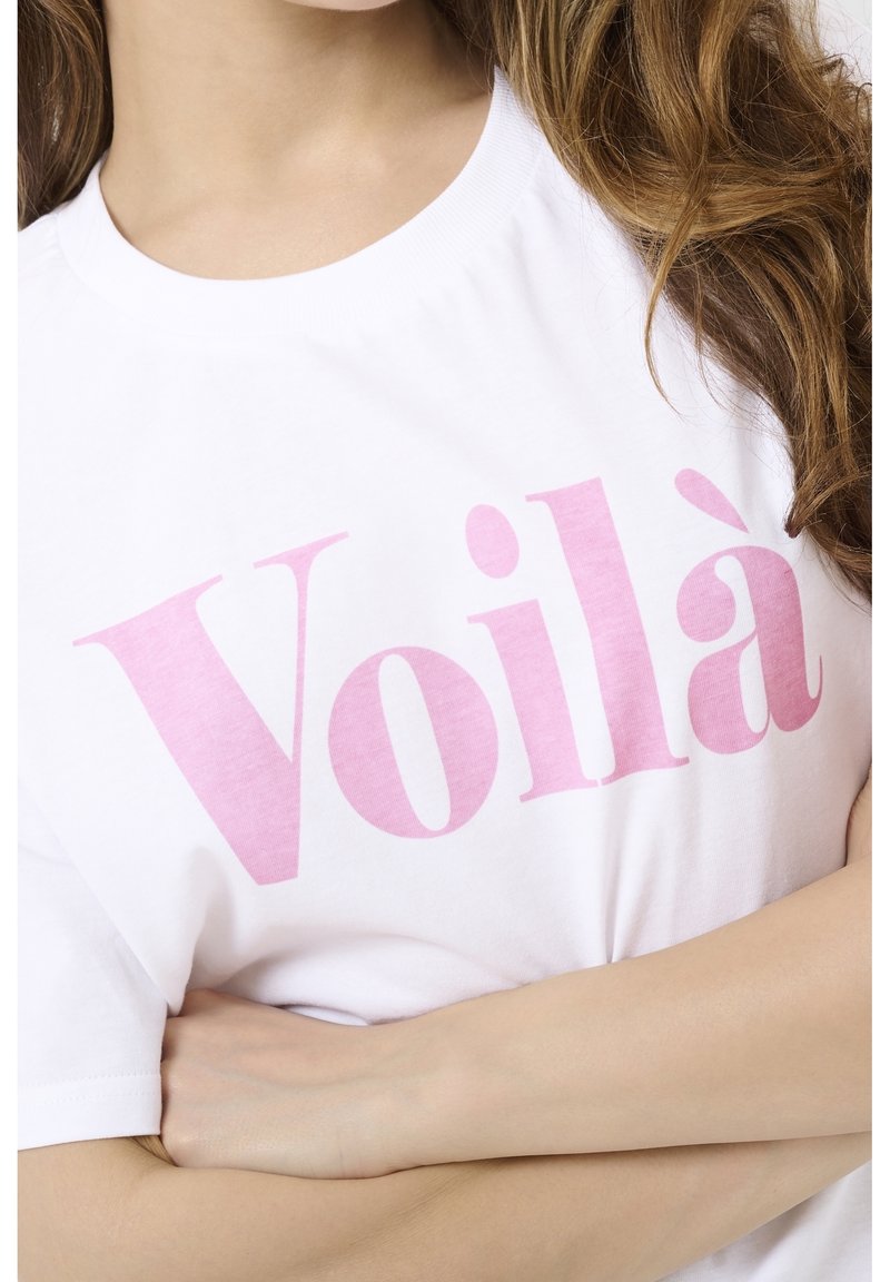 Femme aux longs cheveux portant un t-shirt blanc avec un grand texte rose "Voilà" sur la poitrine, les bras croisés à la taille.