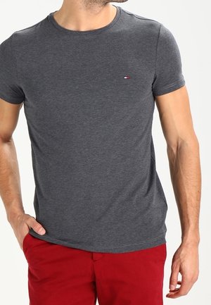 Mann trägt ein schlichtes dunkelgraues Kurzarm-T-Shirt mit kleinem Logo und rote Hose, eine Hand in der Tasche, steht vor einfachem Hintergrund.