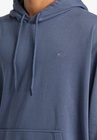 Blue cotton hoodie with a front pocket, adjustable drawstring hood, and a small embroidered logo on the chest. Soft texture.
