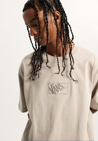 Camiseta de algodón beige con un parche del logo de Vans cosido. El diseño tiene un corte relajado y mangas cortas con una textura suave.