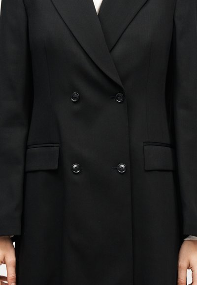 Μαύρο διπλόμπρεστο σακάκι με τέσσερα κουμπιά, τσέπες με καπάκι και λευκό πουκάμισο ορατό στον γιακά και τις μανσέτες.