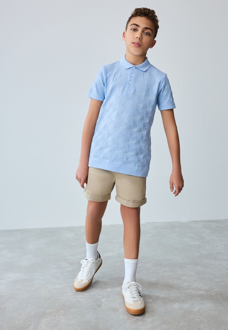 Vaaleansininen, teksturoidulla pinnalla varustettu lyhythihainen pikeepaita, yhdistetty beigen värisiin shortseihin. Valkoiset tennarit kumipohjalla ja käännetyt lahkeet shortseissa.