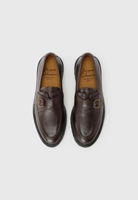 Mocassins en cuir marron avec une finition texturée, dotés d'une boucle décorative et d'une semelle en caoutchouc. L'intérieur présente une semelle intérieure beige avec le nom de la marque.