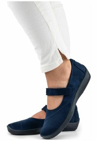 Scarpe in suede blu navy con punta rotonda, una fascia in Velcro sopra l'arco del piede e una suola in gomma nera texturizzata. Indossate con pantaloni bianchi.