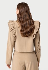 Apkirptas smėlio spalvos švarkas su voliotais pečių akcentais, ilgomis rankovėmis ir tekstūruotu audiniu. Atvirkštinė peržiūra rodo pritaikytą siluetą ir subtilius siūlių elementus.