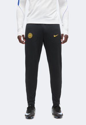 Muž má na sebe čierne športové nohavice Nike s logom Inter Miláno a žltým symbolom Nike, bielu košeľu s dlhým rukávom a čierne kopačky.