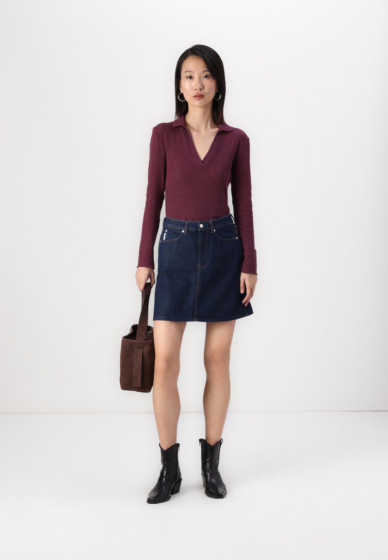 Burgunderfarbener, gerippter Langarmoberteil mit V-Ausschnitt, kombiniert mit einem dunkelblauen Jeans-Minirock. Schwarze Stiefeletten und eine braune Wildleder-Tasche runden das Outfit ab.