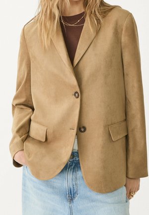 Blazer en daim beige à deux boutons, porté sur un top marron et un jean bleu clair, accessoirisé de colliers et de bagues en or.