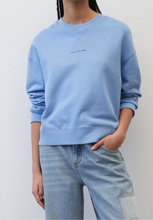Personne portant un sweat-shirt bleu clair à col rond et un jean bleu délavé, main dans la poche, sur fond uni.