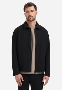 Mann mit Bart, der eine schwarze Zip-Jacke und ein beige Hemd trägt, steht vor einem schlichten weißen Hintergrund.