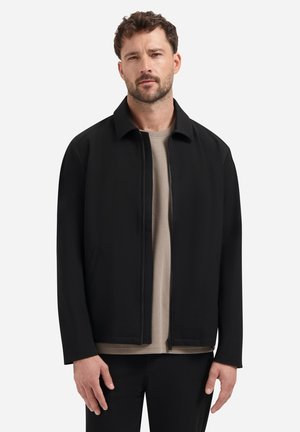 Man met een baard, gekleed in een zwarte bestanden jack en een beige shirt, staande tegen een effen witte achtergrond.