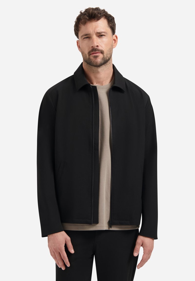 Mann mit Bart, der eine schwarze Zip-Jacke und ein beige Hemd trägt, steht vor einem schlichten weißen Hintergrund.