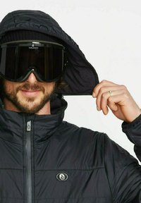 Schwarze isolierte Jacke mit Kapuze, ausgestattet mit einem Reißverschluss und einem Emblem. Die Person trägt große schwarze Skibrillen und eine Strickmütze.