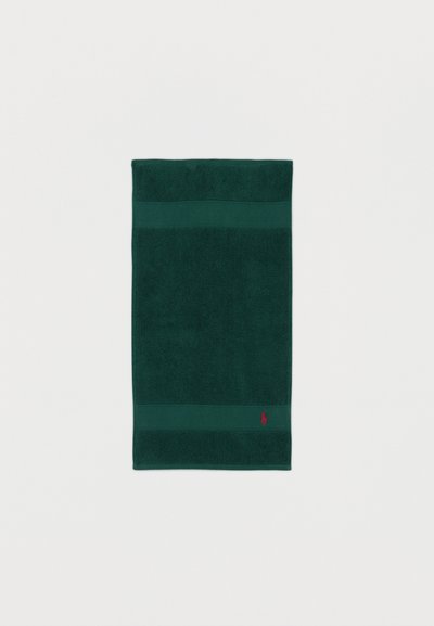 Σκούρα πράσινη ορθογώνια πετσέτα με υφασμένη περίμετρο και ένα μικρό κόκκινο κεντημένο λογότυπο κοντά σε μια γωνία, σε λευκό φόντο.