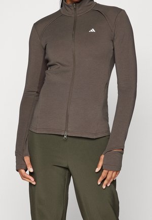 Vrouw draagt een nauwsluitend bruin jasje met ritssluiting en duimgaten, en olijfgroene broek, staand tegen een effen achtergrond.