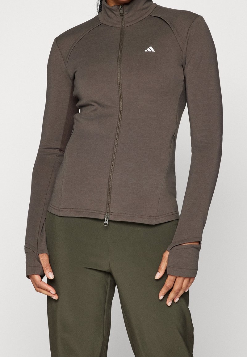 Femme portant une veste marron ajustée à fermeture éclair avec des passe-pouces et un pantalon vert olive, se tenant devant un fond uni.