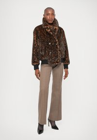 Mujer vestida con una chaqueta de peluche de imitación de leopardo, pantalones de cuadros marrones, medias negras transparentes y zapatos de tacón negros, de pie contra un fondo blanco.