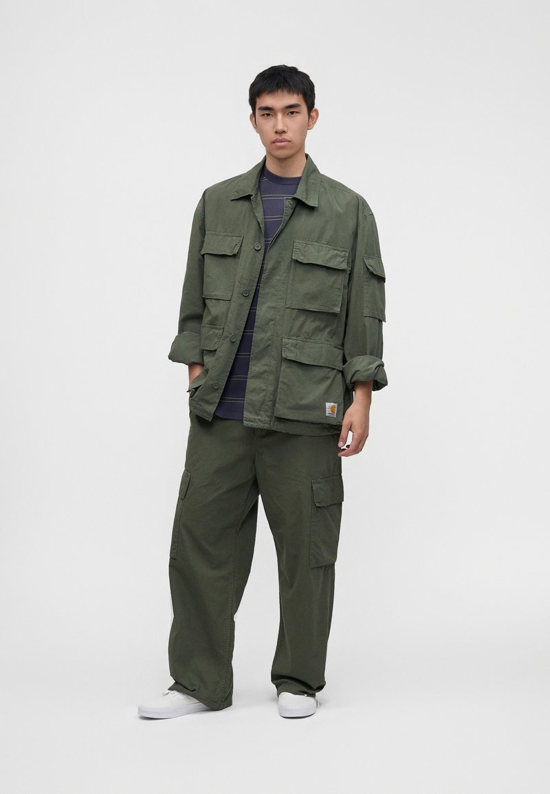 Joven con chaqueta y pantalones cargo verde oliva, camisa azul marino con rayas y zapatillas blancas, de pie con las manos en los bolsillos.