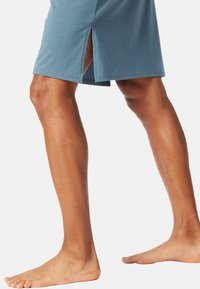 Shorts en coton bleu avec un design à fente latérale, offrant une coupe décontractée et une texture lisse, portés pieds nus.