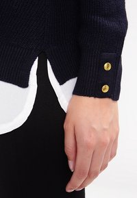 Mörkblå stickad tröja med ribbat mönster, prydd med guldfärgade knappar vid ärmen och en lager-på-lager vit skjortslits som syns undertill.