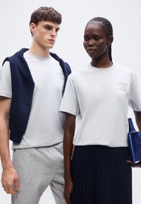 Δύο μοντέλα φορούν λευκά πουκάμισα με κοντά μανίκια· το ένα έχει ένα σκούρο μπλε πουλόβερ περασμένο στους ώμους, ενώ το άλλο κρατά μια υφασμάτινη μπλε τσάντα χειρός.