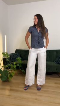 Γυναίκα που στέκεται μέσα σε εσωτερικό χώρο κοντά σε έναν πράσινο καναπέ και φυτά, φοράει μπλε μπλούζα, λευκό παντελόνι με φαρδιά μπατζάκια και μπλε σανδάλια με λουράκια, με τα χέρια στις τσέπες.