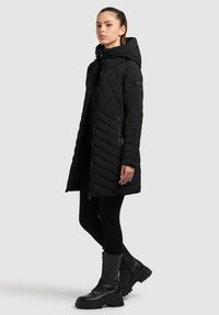Zwarte gewatteerde lange jas met een capuchon, voorzien van diagonale patronen en twee zijritsen. Draagt over zwarte leggings en dikke zwarte laarzen.