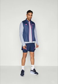 Homme debout portant un gilet de sport bleu marine et rose, une chemise à manches longues claire, un short bleu marine, des chaussettes blanches et des chaussures de course bleu marine sur fond blanc.