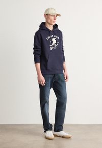 Sudadera azul marino con el logo blanco "GANT USA Ski Club", bolsillo tipo canguro, combinada con jeans azul oscuro y zapatillas blancas. Gorra beige en la cabeza.