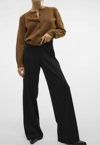 Hnědý pletený cardigan s knoflíky, zkombinovaný s černými kalhotami s širokými nohavicemi a elegantními černými botami. Hladká textura a přizpůsobený střih.