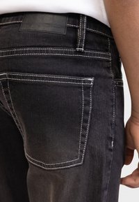 Μαύρα τζιν από τζιν ύφασμα με λευκή ραφή, δερμάτινο ετικέτα στη ζώνη μέσης και κοντινή άποψη στην πίσω τσέπη και το χέρι.