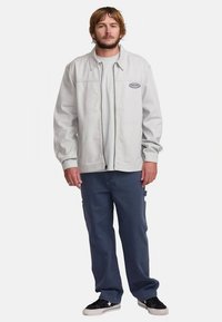 Light gray zip-up jacket with front pockets, worn over a gray T-shirt. Paired with dark blue loose-fit pants and black sneakers.