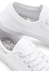 White canvas sneakers with round toe, lace-up design, and textured rubber sole. Interior features a patterned insole. Logo on side.