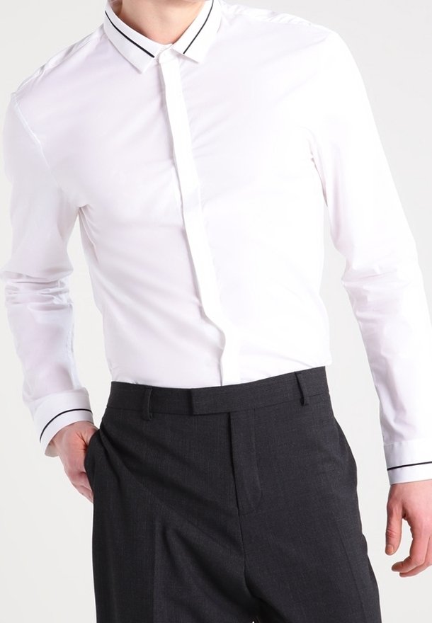 Homme portant une chemise blanche avec un liseré noir sur le col et les poignets, et un pantalon noir, debout avec une main dans la poche.