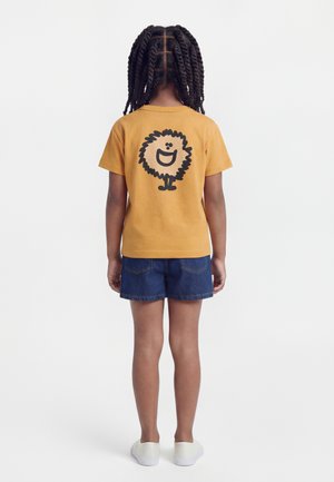 Otrok s kitkami, ki nosi gorčično majico z nasmejanim črnim risanim likom na hrbtni strani, kratke jeans hlače in bele natikače, stoji.