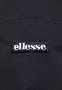 Černá textilie s viditelným prošíváním; obsahuje obdélníkovou aplikaci s bílým vyšitým názvem značky "ellesse."