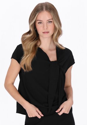 Hosszú, hullámos szőke hajú nő, fekete, rövid ujjú, elöl megkötős felsőt visel, fehér háttér előtt áll.