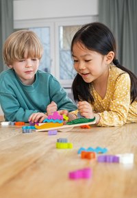 Twee kinderen spelen met kleurrijke, gestructureerde bouwblokken van zacht materiaal, uitgestald op een houten oppervlakte. De vormen omvatten golven en boten.