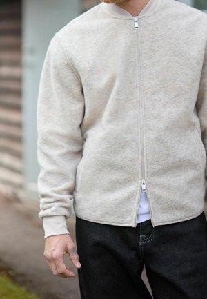 Homme portant une veste beige zippée et un pantalon noir, debout à l'extérieur avec une main partiellement visible montrant de petits tatouages.