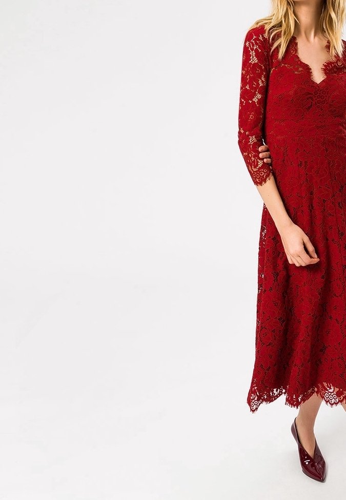 Femme en robe midi rouge en dentelle à manches trois-quarts et chaussures bordeaux pointues, debout devant un fond blanc uni.