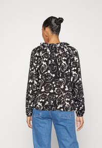 Blusa nera con motivo floreale bianco. Presenta un collo arricciato e maniche lunghe con polsini elasticizzati. Indossata con jeans in denim blu chiaro.